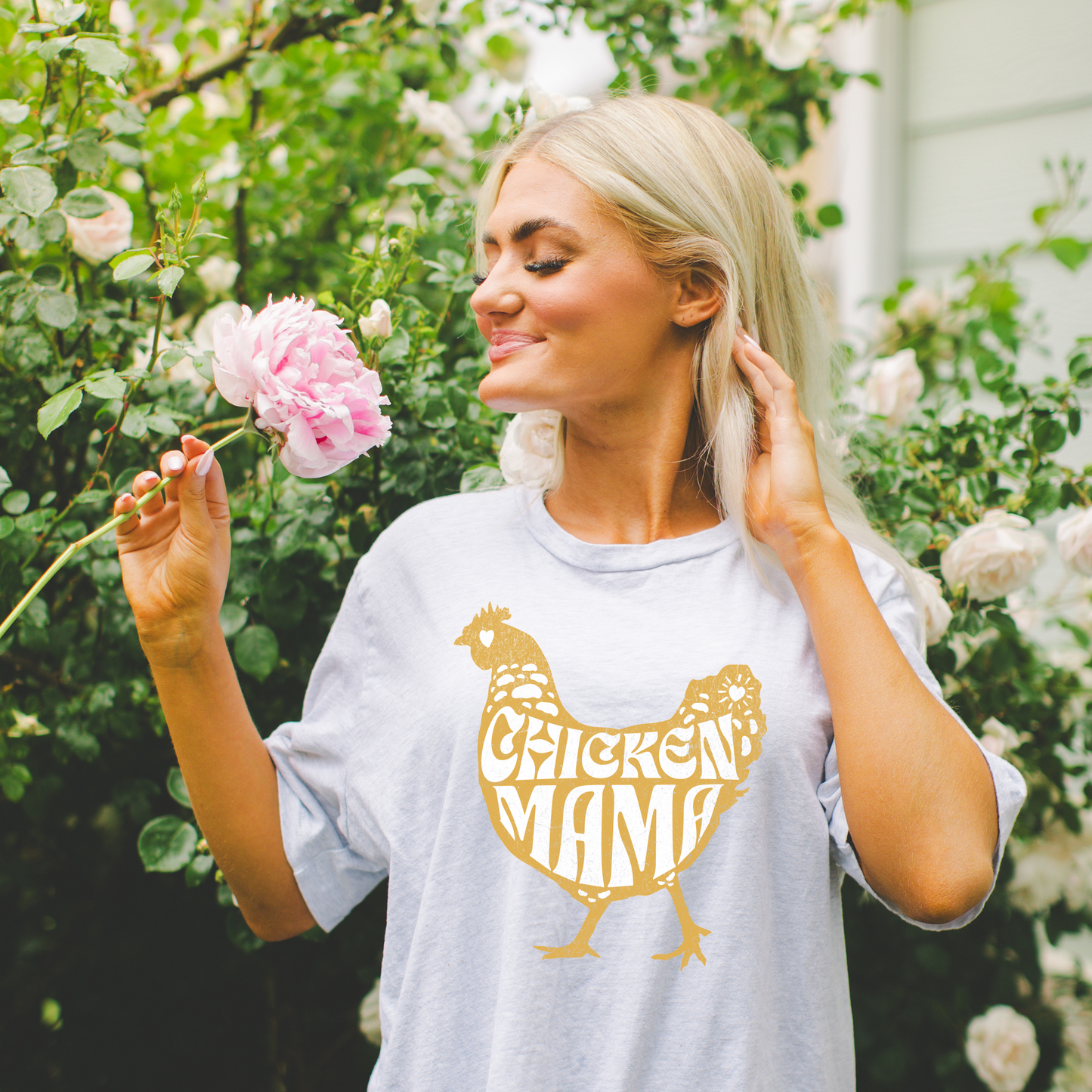 A woman smelling a pink flower, wearing a gray t-shirt with 'Chicken Mama' text in a chicken shape design.