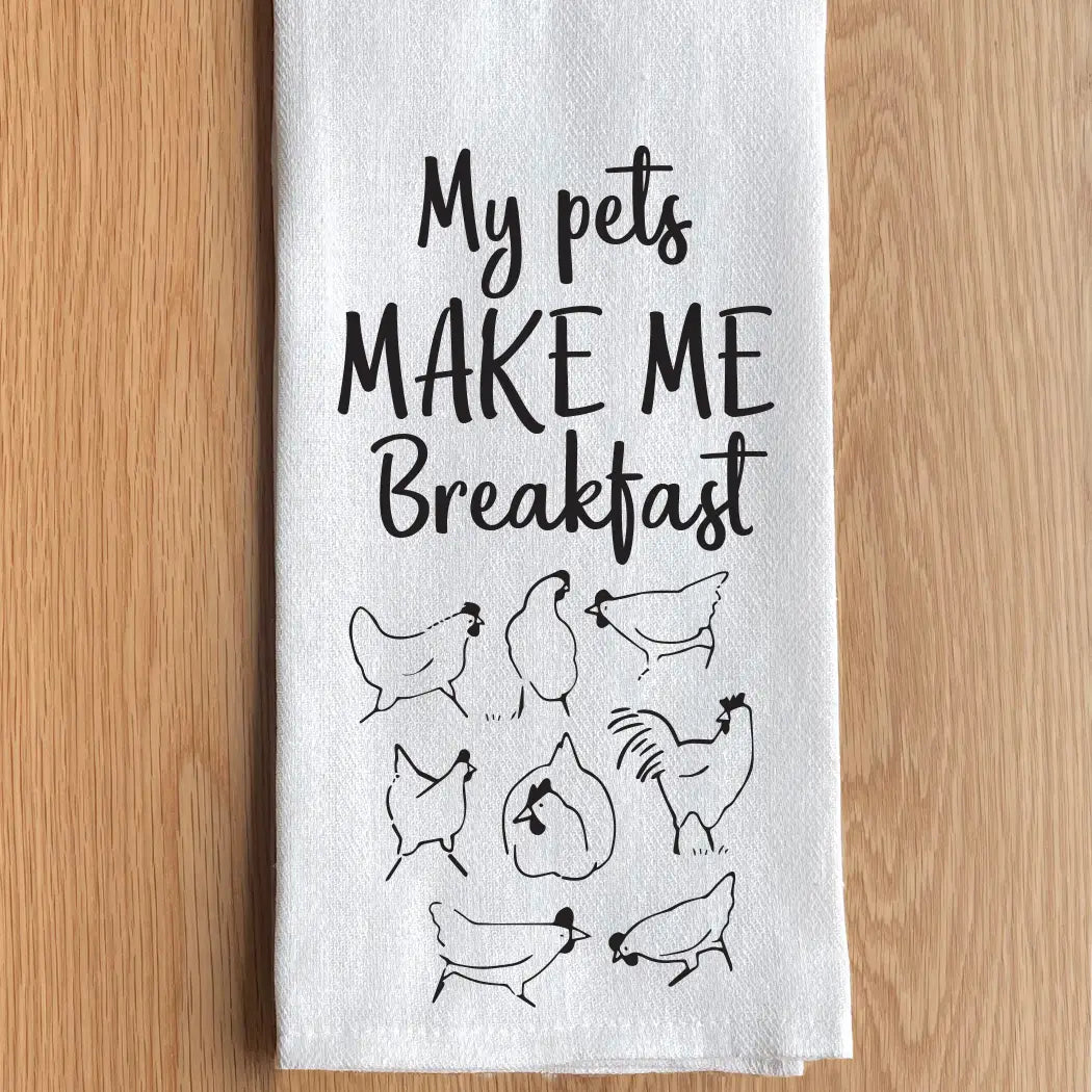 Kitchen towel with text 'My pets MAKE ME Breakfast' and chicken illustrations on a wooden surface